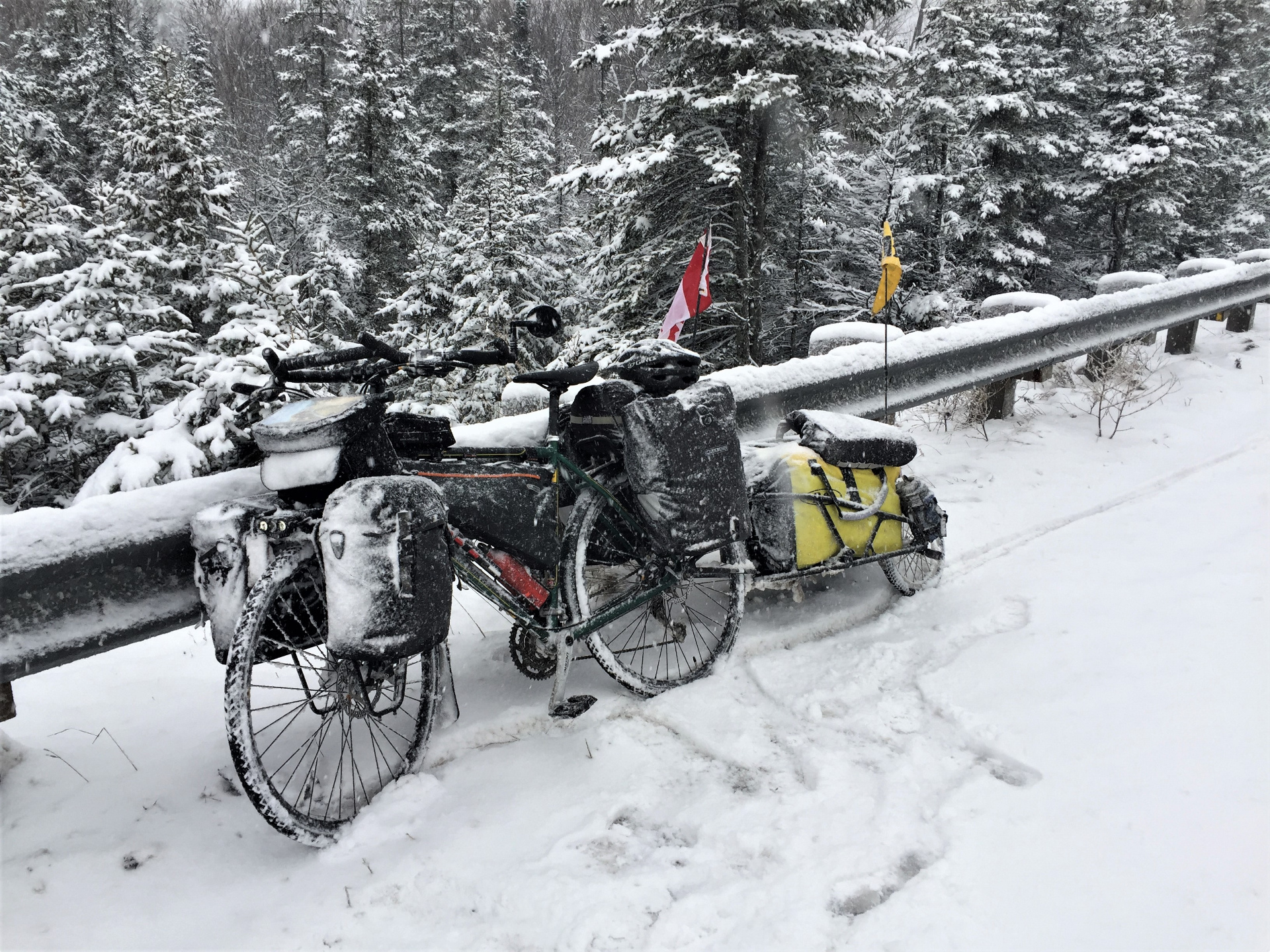A loaded touring bicycle with a Canadian flag, covered in snow, parked beside a guardrail in a snowy forest.