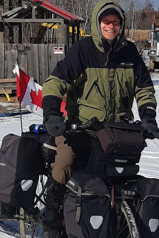 The author bundled in a winter jacket, standing behind a fully loaded touring bicycle with a Canadian flag, snow on the ground.